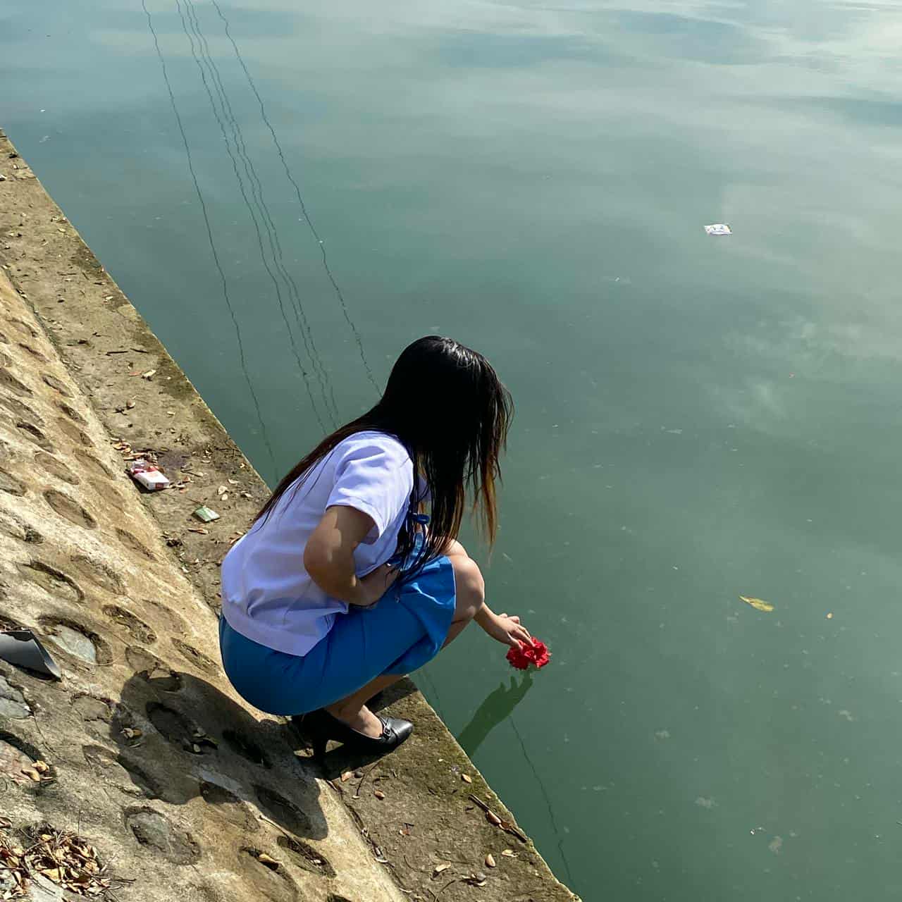 Young woman stands beside a river holding a green flowering plant, under a vivid blue sky with scattered clouds. The scene is outdoors, peaceful, and relaxed. The image has enhanced contrast and a pronounced 3D tilt effect.