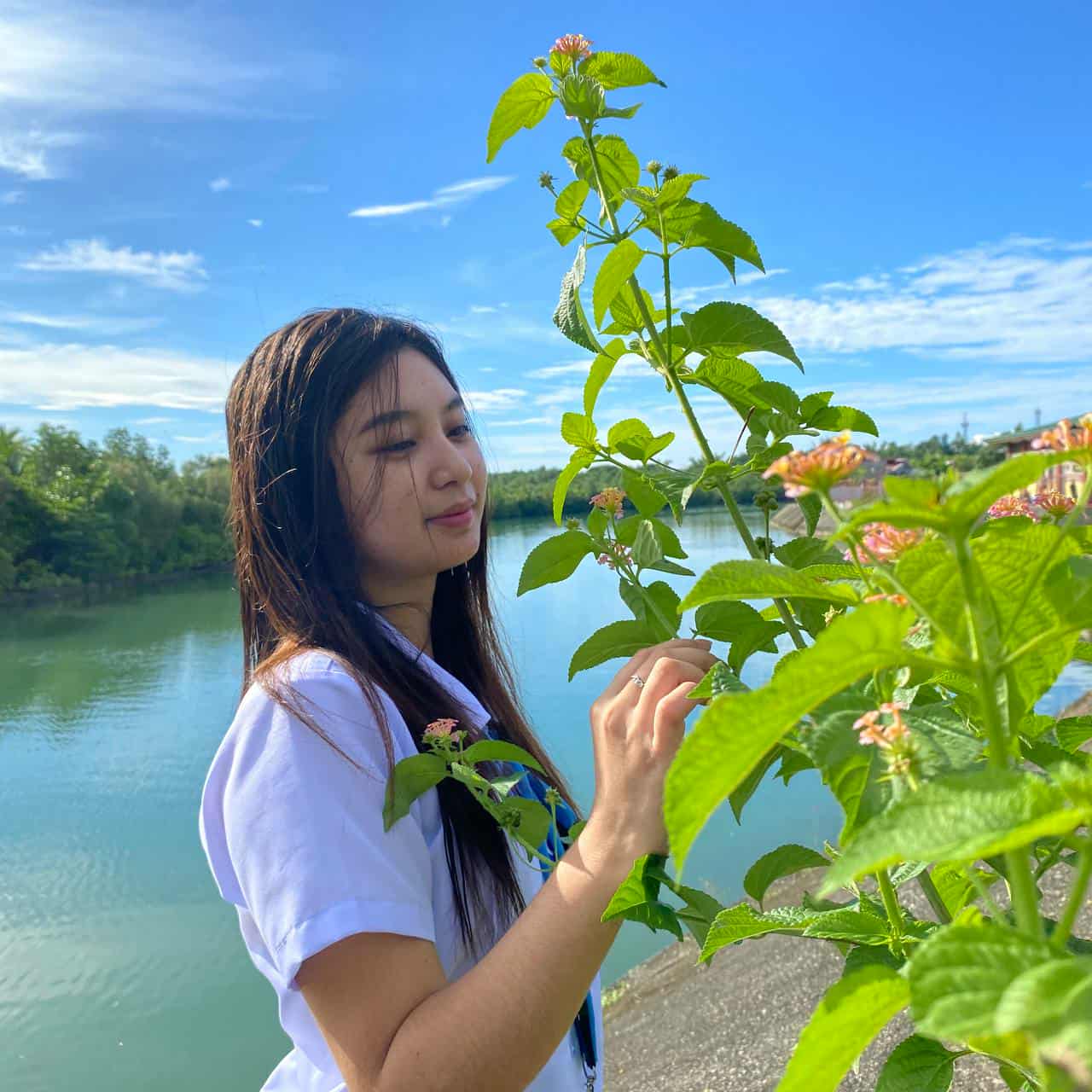 Young woman stands beside a river holding a green flowering plant, under a vivid blue sky with scattered clouds. The scene is outdoors, peaceful, and relaxed. The image has enhanced contrast and a pronounced 3D tilt effect.